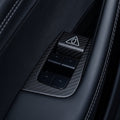 Close-up of four black window switches inside a carbon-weave surround with a triangular door-release cover at one end.